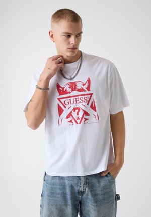 Junger Mann mit kurzen blonden Haaren, der ein weißes T-Shirt mit rotem grafischem Design und verblasste blaue Jeans trägt, schaut zur Seite.