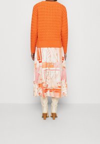 Pomarańczowy sweter o teksturowanym splocie zestawiony z luźną, wzorzystą spódnicą w odcieniach brzoskwiniowym, różowym i białym, do której dodano beżowe, sięgające kolan botki.