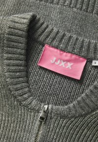 Pilkas megztinis su guliu tekstūra, su rožiniu audinio žymeniu "JJXX" ir sidabriniu užtrauktuku ant kaklo.