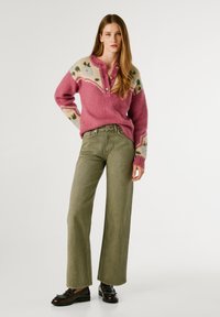Suéter de punto rosa con patrón decorativo y detalle de botones, combinado con pantalones de pierna ancha en verde oliva y mocasines negros con tachuelas.