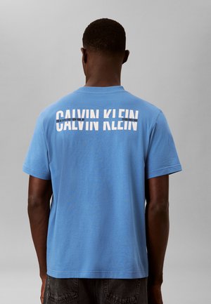 Mand iført en lyseblå Calvin Klein T-shirt med hvidt logotekst på øverste del af ryggen, stående mod en ensfarvet grå baggrund.