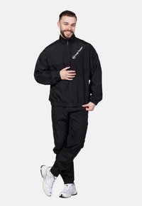 Schwarzer leichter Trainingsanzug mit einer Jacke mit Reißverschluss und elastischen Bündchen, mit einem Logo auf der Brust. Das Modell trägt weiße Sneakers.