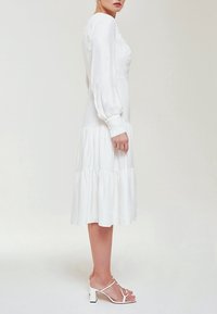 Biała sukienka z długim rękawem i warstwową spódnicą, zdobiona haftem kwiatowym oraz mankietami. W parze z białymi sandałami na obcasie, charakteryzującymi się minimalistycznym wzornictwem.