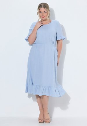 Frau mit blonden Haaren steht und trägt ein hellblaues knielanges Kleid mit Rüschen an den kurzen Ärmeln und am Saum, kombiniert mit hautfarbenen Pumps mit Knöchelriemen.