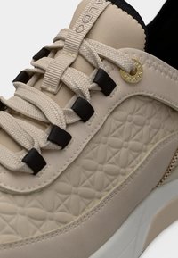 Baskets beiges avec un panneau latéral embossé à motifs floraux, des lacets beiges passés dans des œillets noirs, et un détail métallique doré avec le logo sur le trou de lacet.