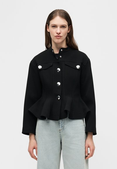 Γυναίκα που φοράει μαύρο εφαρμοστό σακάκι με ασημένια κουμπιά και βολάν στο τελείωμα, σε συνδυασμό με ανοιχτόχρωμα μπλε τζιν, στέκεται μπροστά σε απλό φόντο.