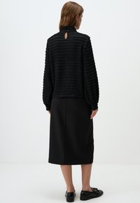 Zwarte tekstuurblouse met een hoge hals en sleutelgatdetail, gecombineerd met een zwarte midi-rok. Het schoeisel heeft glanzende zwarte accenten.