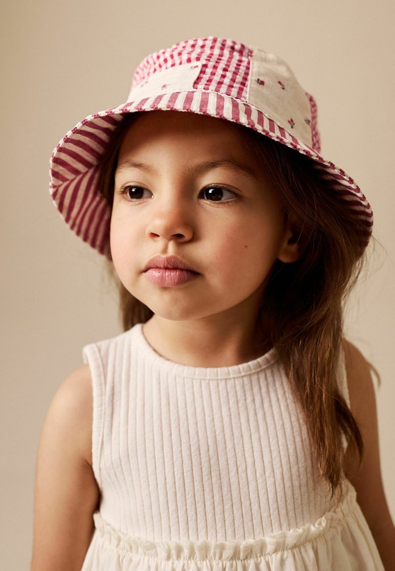 Niña joven con pelo largo y castaño que lleva un sombrero bucket rojo y blanco a rayas y un vestido blanco sin mangas, mirando ligeramente hacia un lado.