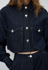 Tummansininen lyhyt takki, jossa on hopeiset nappikiinnitykset, kaksi rintataskua ja näkyvät ompelutiedot. Yhdistetty vastaaviin korkeavyötäröisiin housuihin.