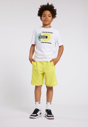 T-shirt blanc avec un design de logo "BOSS" coloré, associé à un short jaune vif. Les chaussures comprennent des baskets noir et blanc avec des chaussettes à motifs.
