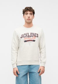 Νέος άνδρας που φοράει ελαφρύ μπεζ φούτερ με το λογότυπο "JACK & JONES" και μπλε τζιν, στέκεται μπροστά σε απλό λευκό φόντο.