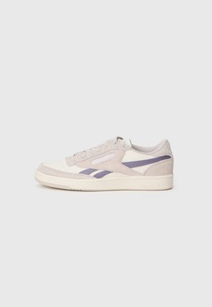 Baskets basses Reebok beige et blanches avec des bandes latérales violettes, dessus en suède et cuir, et fermeture à lacets sur un fond clair.