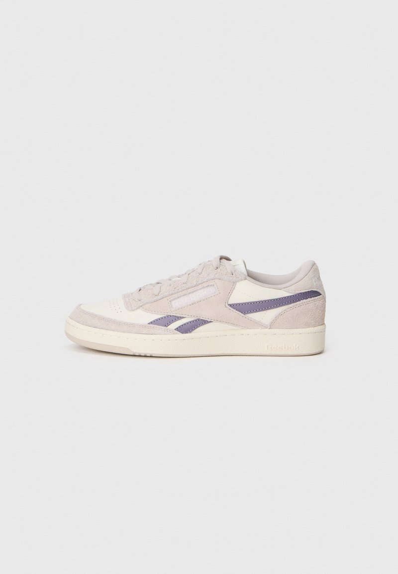 Baskets basses Reebok beige et blanches avec des bandes latérales violettes, dessus en suède et cuir, et fermeture à lacets sur un fond clair.