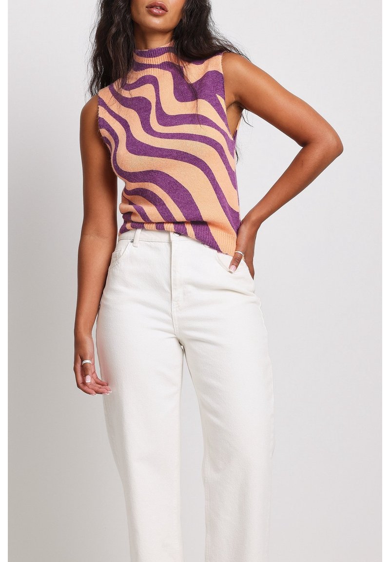 Pull sans manches violet et pêche avec motif ondulé, texture tricotée. Associé à un pantalon blanc taille haute, créant une tenue décontractée.