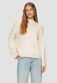 Beiger Strickpullover mit lockerer Passform, überschnittenen Schultern und geripptem Saum, kombiniert mit hellblauen Jeans. Weiche Textur und warmes Material.