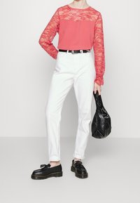 Koraalkleurige lange mouwen top met kantdetails, witte high-waisted broek, zwarte riem, zwarte handtas en zwarte schoenen met grove zolen.
