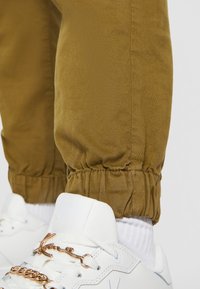 Pantalon de jogging marron retroussé au-dessus des chaussettes blanches et baskets blanches avec chaîne en or et détail signature sur les lacets.
