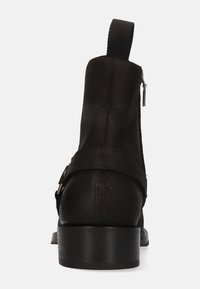 Bota de cuero negro para tobillo con textura suave, lengüeta trasera para tirar, cremallera lateral y tacón plano. Cuenta con una correa decorativa que cruza el tobillo.