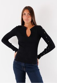Top nero a maniche lunghe a costine con scollo a V, dettagli metallici sulle spalle e sui polsini, vestibilità aderente. Indossato con jeans blu.