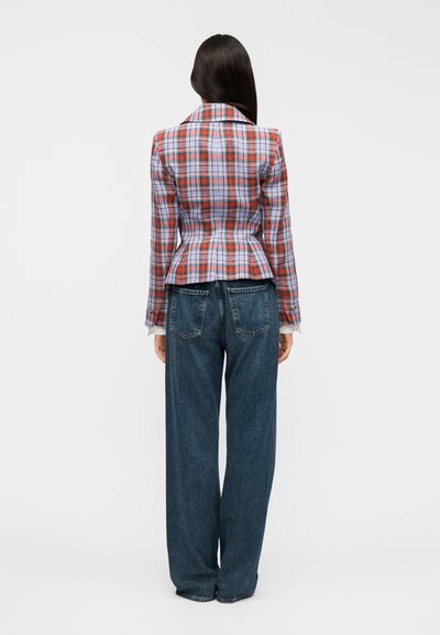 Καρό σακάκι σε κόκκινο, μπλε και λευκό με εφαρμοστή σιλουέτα, μέση με πεπλούμ και γιακάδες με σχισμές, συνδυασμένο με φαρδιά μπλε τζιν παντελόνια.