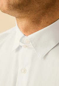 Weißes Hemd mit spitzem Kragen, das eine glatte Textur, zwei Knopfverschlüsse und einen dezenten Glanz aufweist. Nahaufnahme des Kragenbereichs.