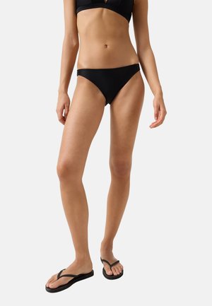 Kobieta w czarnych bikini i czarnych japonkach, stojąca w swobodnej pozie na jednolitym jasnym tle.