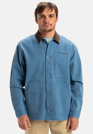 Blue canvas jacket with a brown collar, featuring front buttons, two side pockets, and a chest pocket. Simplistic design, sturdy texture.