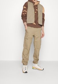 Beiget työpöksyt sivutaskuilla, ruskea huppari, jossa leikkauskuvioita, ja vaaleanbeige liivi. Kengät ovat valkoiset, joissa värikkäitä yksityiskohtia.