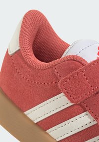 Červené semišové tenisky s bielymi koženými pruhmi a suchým zipsom. Majú textúrované vypchávky okolo členku. Gumová podrážka.