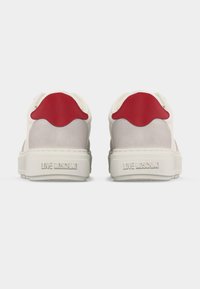 Ζευγάρι λευκά και μπεζ αθλητικά παπούτσια με κόκκινες φτέρνες και το λογότυπο "Love Moschino" χαραγμένο στη φτέρνα, πάνω σε λευκό φόντο.