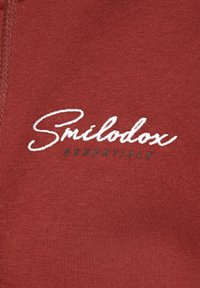 Un tessuto color ruggine presenta un testo in corsivo bianco che recita "Smilodon" con "ESSENTIALS" in un font scuro più piccolo sottostante. Superficie morbida e texturizzata.