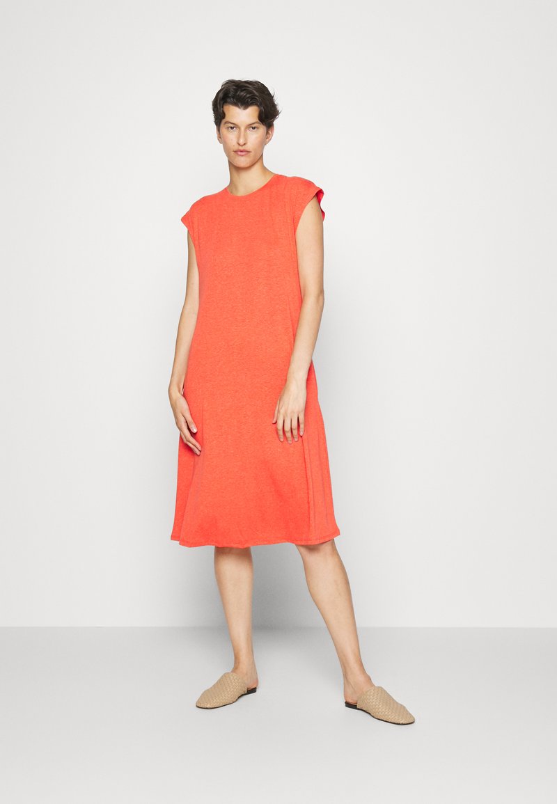 Vestido coral claro de mangas curtas, corte solto, até ao joelho. Tecido suave e texturizado com um design simples. Usado com sapatos slip-on castanhos.