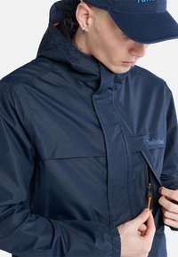 Giacca impermeabile blu navy con colletto alto, tasca con zip e polsini elasticizzati. Presenta un logo sul petto e una zip arancione a contrasto.