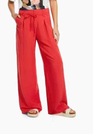 Personne portant un pantalon rouge ample avec une taille à cordon, associé à des sandales blanches à plateforme, debout avec une main dans la poche.
