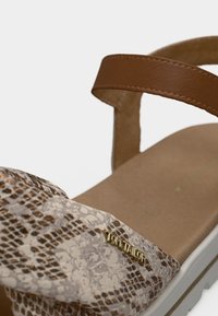 Sandales à brides en cuir marron avec une tige synthétique beige à motifs et un détail logo doré. La semelle est blanche et texturée pour une meilleure adhérence.