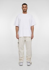 Maglietta oversize bianca in cotone, abbinata a pantaloni cargo beige chiaro con tasche laterali e dettagli neri. Sneakers grigie completano l'outfit.