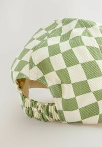 Zielono-kremowa czapka w kratę z materiału sztruksowego, z zebranym elastycznym tyłem umożliwiającym regulację dopasowania. Teksturowany materiał z wyraźnymi wzorami.