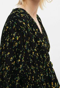 Robe noire à fleurs avec des fleurs jaunes et vertes, texture plissée, encolure en V et manches amples, créant une silhouette décontractée.