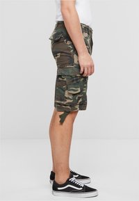 Maskovací cargo kraťasy vyrobené z bavlněného twillu, s bočními kapsami, knoflíkovými detaily a ohrnutými lemy. Nosené s černobílými teniskami.