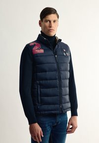 Chaleco acolchado azul marino con costuras horizontales, cierre con cremallera en la parte delantera y dos bolsillos laterales. Presenta un emblema rojo y blanco en el hombro izquierdo.