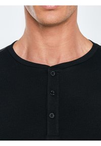 Zbliżenie na szyję i górną część klatki piersiowej mężczyzny noszącego czarną prążkowaną koszulkę henley z trzema czarnymi guzikami.