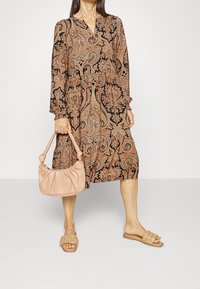 Vestido de mangas largas con estampado paisley en tonos cálidos y escote en V. La modelo lleva un bolso suave de color rosa claro y sandalias de tiras beiges.