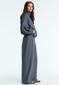 Kobieta ubrana w długą, luźną sukienkę w paski z długimi rękawami i wysokim kołnierzem, stojąca z rękami w kieszeniach na jednolitym tle.