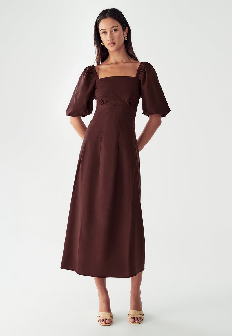 Rjava dolga obleka s pufastimi rokavi in kvadratnim izrezom, gladka tekstura tkanine in oprijet pas, v kombinaciji s svetlo peščenimi sandali na platformi.