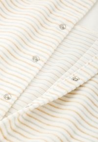 Tissu rayé en crème et beige, présentant une surface délicatement texturée. Des boutons-pression sont visibles le long du bord d'un vêtement replié.