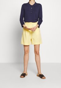 Marinblå långärmad polo med tre knappar, i kombination med gula shorts och bruna halksandaler. Slätt tyg med avslappnad passform.