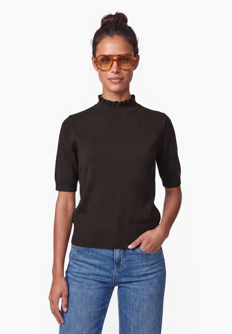 Femme aux cheveux foncés en chignon portant de grandes lunettes teintées ambre, un haut noir à manches courtes et un jean bleu, une main dans la poche.
