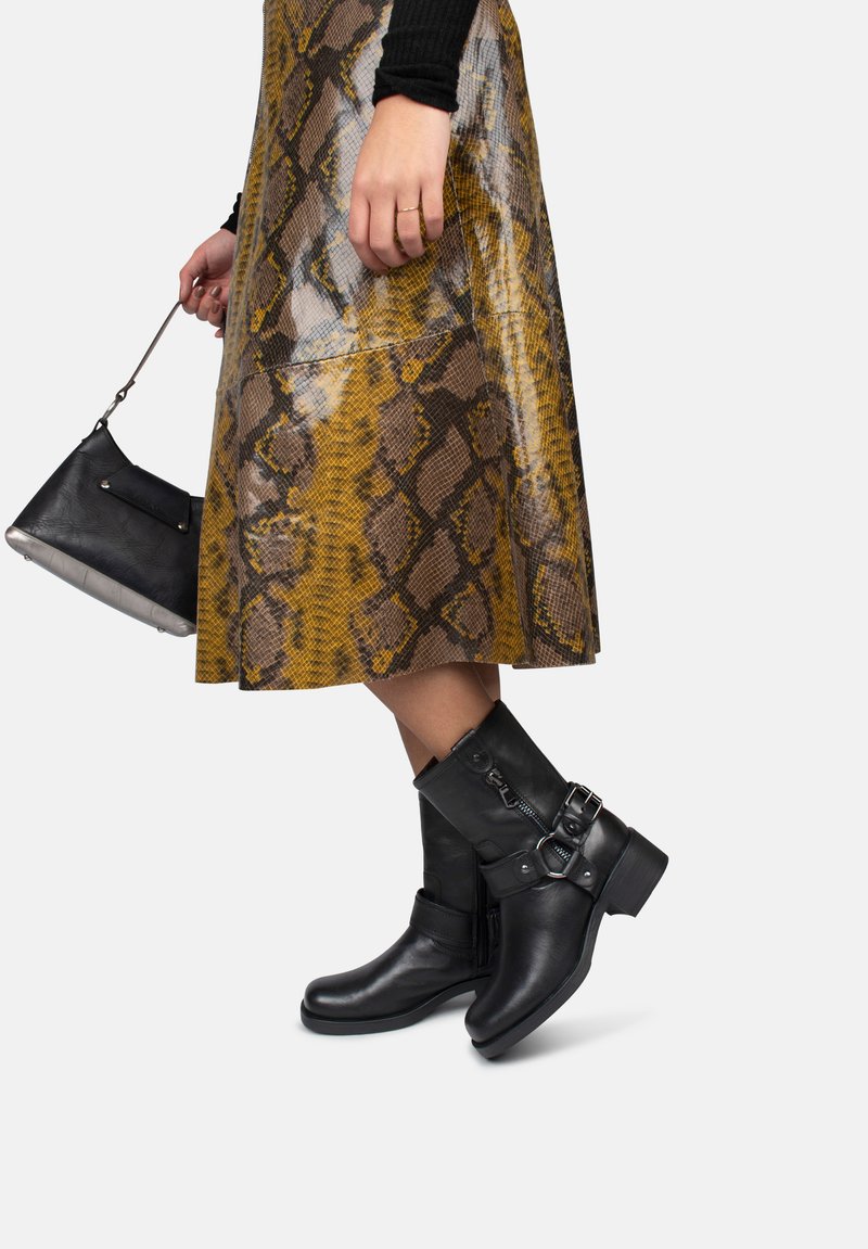 Botas de cuero negro hasta el tobillo con un tacón grueso, detalles de hebilla y cremalleras laterales. Llevadas con una falda amarilla y marrón con estampado de piel de serpiente.