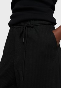 Pantalones negros con un sutil patrón de rayas finas, ajuste slim, cinturilla elástica con cordón, bolsillos laterales y detalle de logo bordado.