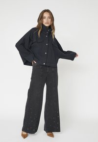 Übergroße, schwarz gestreifte Jacke mit Druckknöpfen und zwei Fronttaschen, kombiniert mit schwarzen weiten Jeans, die am Saum Ösenakzente aufweisen.